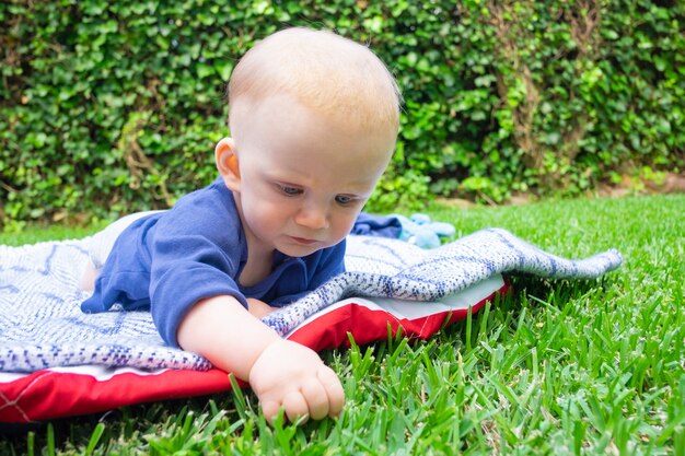 Śliczne rudowłose dziecko w niebieskiej koszuli dotykając trawy w parku i leżąc na brzuchu. Piękna mama ogląda niemowlę w parku i uśmiecha się. Letni czas dla rodziny