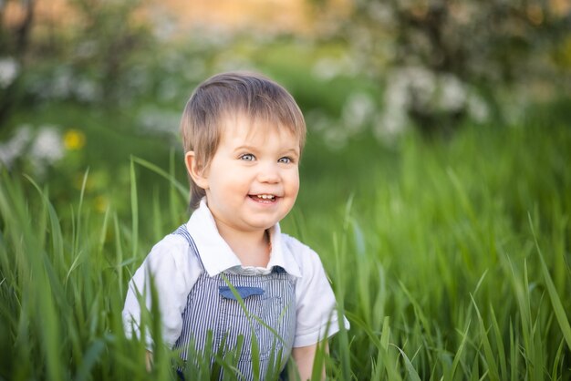 Śliczny chłopiec w modnych ubraniach z niebieskimi oczami bawi się i skacze w wysokiej trawie w dużym zielonym kwitnącym ogrodzie