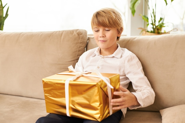 Śliczny kaukaski nastolatek siedzi na kanapie z prezentem noworocznym na kolanach. Przystojny chłopak gotowy do otwarcia złotego pudełka z prezentem w nim, mając ciekawy przewidywany wyraz twarzy, uśmiechając się