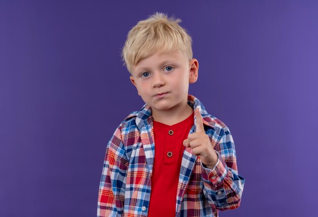 Śliczny mały chłopiec z blond włosami w koszuli w kratkę pokazujący palec wskazujący, patrząc na fioletową ścianę