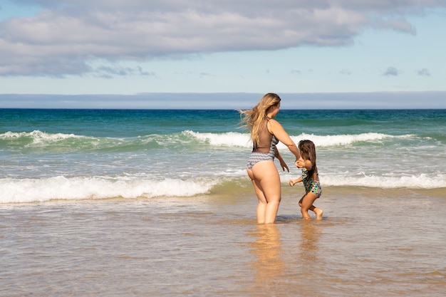 Słodka młoda mama i córeczka stoją po kostkę w wodzie morskiej, spędzając wolny czas na plaży nad oceanem