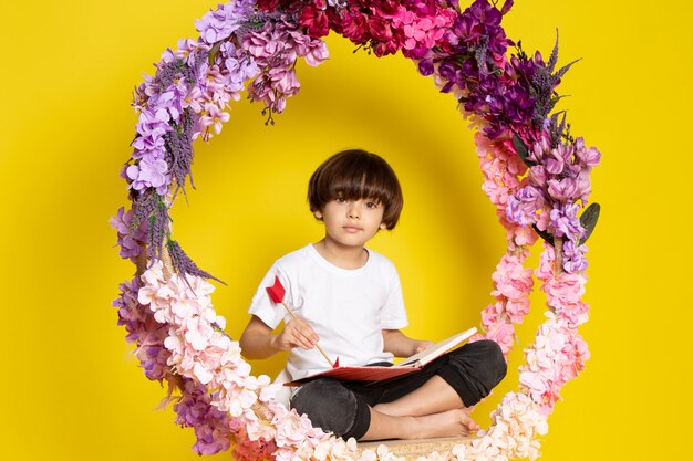 Słodki, uroczy, słodki chłopiec w widoku z przodu w białej koszulce, siedzący na kwiatku, stał na żółtej podłodze