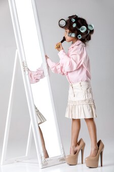 Słodkie małe dziecko dziewczynka w lokówki do włosów bawi się ubraniami i butami matki przed lustrem.