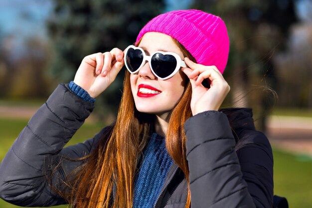 Słoneczny wiosenny portret szczęśliwej wesołej uśmiechniętej imbirowej kobiety pozującej w parku, ciesz się słonecznym dniem, jasnym punkowym kapeluszem hipster, serdecznymi okularami przeciwsłonecznymi, czerwonymi ustami, ciepłą parką, pozytywnym nastrojem.
