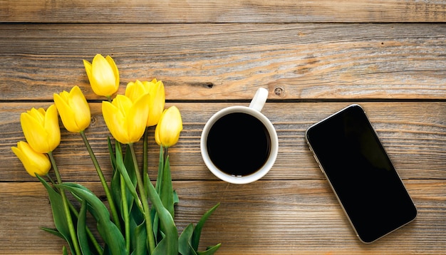 Bezpłatne zdjęcie smartfon filiżankę kawy bukiet żółtych tulipanów na drewnianym tle
