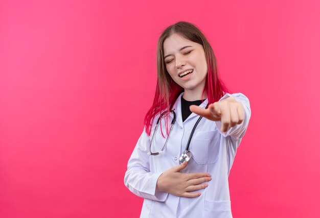 Śmiejąc się młoda kobieta lekarz noszenie stetoskop medycznej sukni pokazującej gest na różowej ścianie na białym tle
