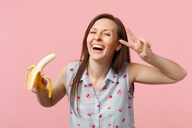 Śmiejąc się młoda kobieta w letnie ubrania pokazując znak zwycięstwa trzymając świeżych dojrzałych owoców bananów na białym tle na różowym tle ściany pastelowe. żywy styl życia ludzi, koncepcja wypoczynku relaksu. makieta miejsca na kopię.