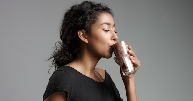 Bezpłatne zdjęcie Śmiejąca się atrakcyjna dziewczyna z bliskiego wschodu w zrelaksowanej czarnej bluzce pijąca kawę mocca z gorącą czekoladą z wysokiej szklanki odizolowanej na szaro