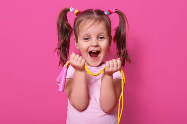 Śmiejąca się śliczna dziewczyna nosi różową koszulę, stoi na różowo, trzyma w rękach jasną skakankę. Szczęśliwe dziecko z otwartymi ustami lubi bawić się nową skakanką. Koncepcja dzieciństwa.
