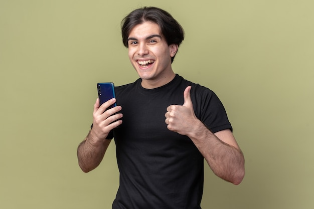 Śmiejący się młody przystojny facet na sobie czarną koszulkę trzymając telefon pokazując kciuk do góry na białym tle na oliwkowej ścianie
