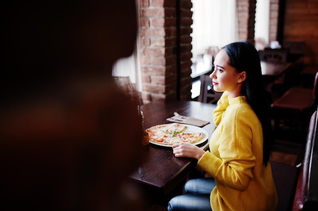 Śmieszna brunetka dziewczyna w żółtym swetrze je pizzy w restauracji
