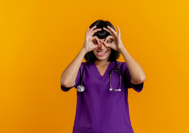 Śmieszna młoda brunetka lekarka w mundurze ze stetoskopem wystaje język