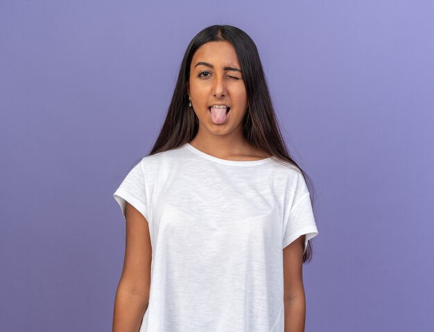 Śmieszna młoda dziewczyna w białej koszulce patrząca na aparat wystający język i mrugający, stojący na niebieskim tle