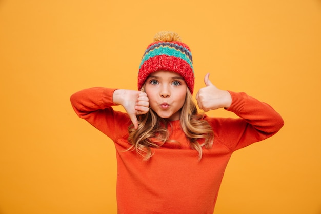 Śmieszna młoda dziewczyna w pulowerze i kapeluszu pokazuje kciuk w górę i na dół podczas gdy patrzejący kamerę nad kolorem żółtym