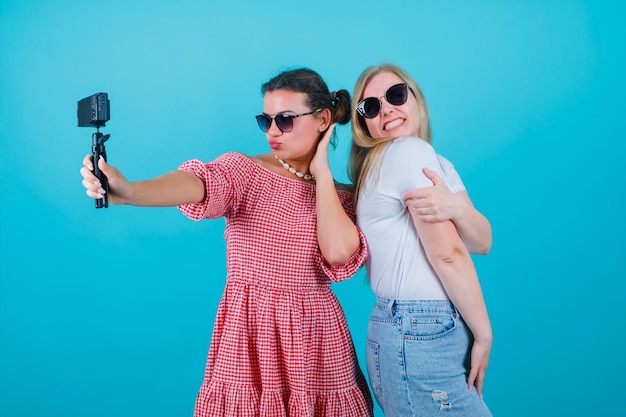 Śmieszne Dwie Dziewczyny Robią Sobie Selfie Swoim Mini Aparatem, Pokazując Różne Pozy Na Niebieskim Tle