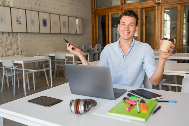 Bezpłatne zdjęcie Śmieszne uśmiechnięty szczęśliwy młody człowiek siedzi w współpracującym pokoju biurowym, pracując na laptopie