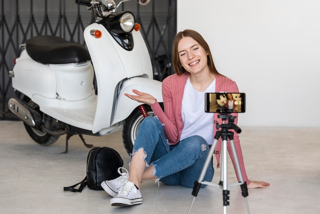 Smiley młoda blogerka nagrywająca się siedząca obok motocykla
