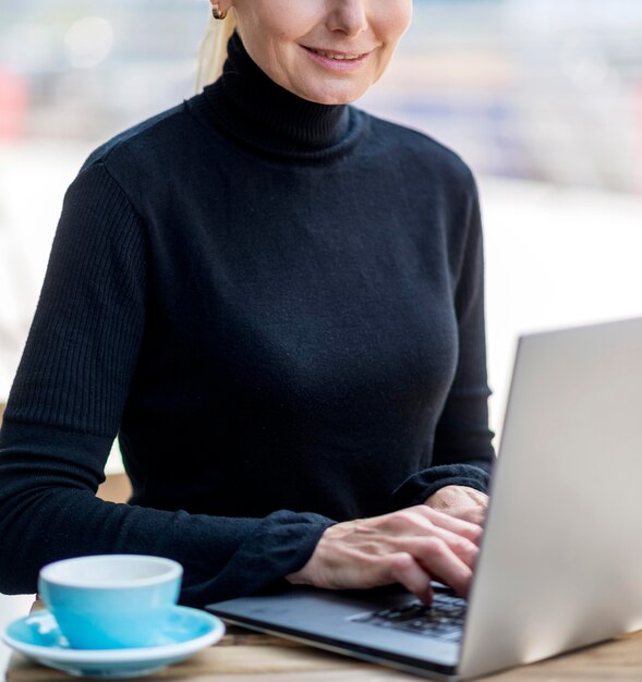 Smiley starsza kobieta pije kawę na zewnątrz podczas pracy na laptopie