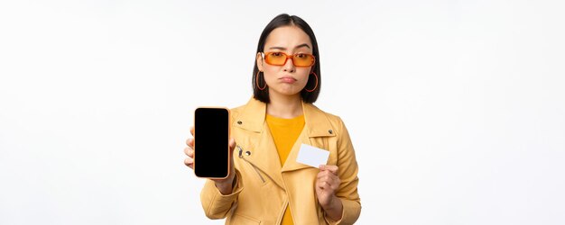 Smutna Azjatycka dziewczyna w okularach przeciwsłonecznych pokazująca kartę kredytową z interfejsem aplikacji smartfona, wyglądającą na rozczarowaną stoisko