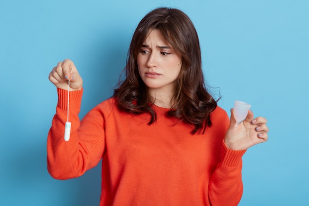 Smutna ciemnowłosa kobieta ubrana w pomarańczowy sweter, trzymając w rękach produkty higieniczne dla kobiet