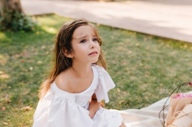 Smutna dziewczyna o jasnobrązowych włosach ma zamiar płakać siedząc na kocu obok alei. Zewnątrz portret nieszczęśliwego dziecka patrząc oczami pełnymi łez w parku.