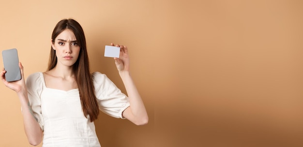 Bezpłatne zdjęcie smutna i rozczarowana dziewczyna pokazująca pusty ekran smartfona i plastikową kartę kredytową narzekającą i fr