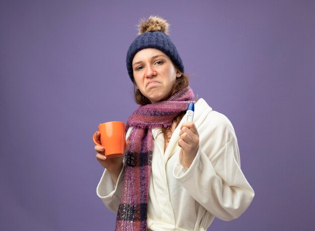 Smutna młoda chora dziewczyna ubrana w białą szatę i czapkę zimową z szalikiem, trzymając filiżankę herbaty z termometrem na fioletowym tle
