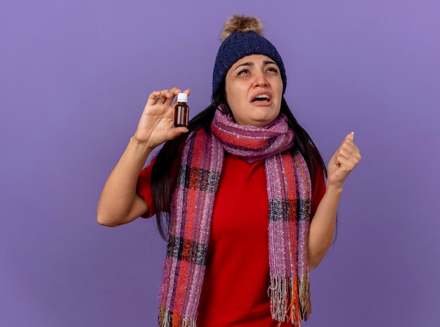 Bezpłatne zdjęcie smutna młoda kaukaska chora dziewczyna w czapce zimowej i szaliku trzymająca lekarstwo w szkle patrząc w górę zaciskającą pięść odizolowaną na fioletowym tle z miejscem na kopię