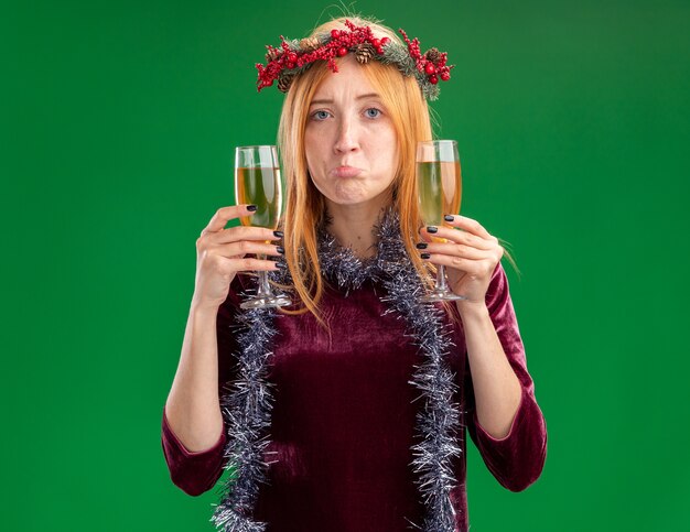 Smutna młoda piękna dziewczyna ubrana w czerwoną sukienkę z wieniec i garland na szyi, trzymając kieliszek szampana na białym tle na zielonym tle