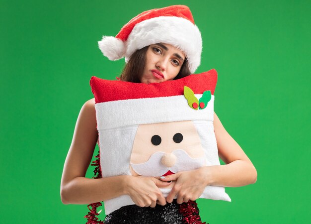 Smutna młoda piękna dziewczyna ubrana w świąteczny kapelusz z girlandą na szyi, trzymając świąteczną poduszkę na białym tle na zielonym tle