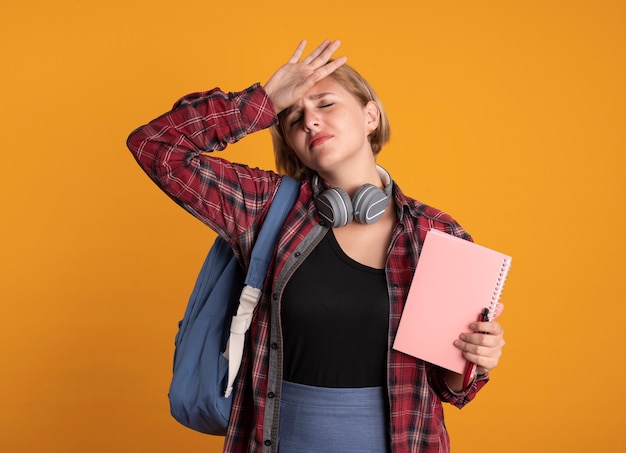 Smutna młoda słowiańska studentka ze słuchawkami w plecaku kładzie rękę na czole trzyma notatnik i długopis