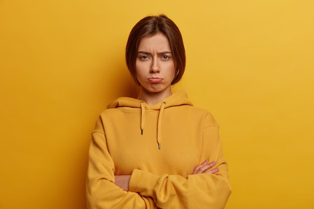 Bezpłatne zdjęcie smutna zdradzona nieszczęśliwa kobieta stoi z założonymi rękami, chce płakać, czuje się urażona i samotna, nosi bluzę, modelki na żółtej ścianie, torebka dolna warga, zastanawia się nad poważnym problemem