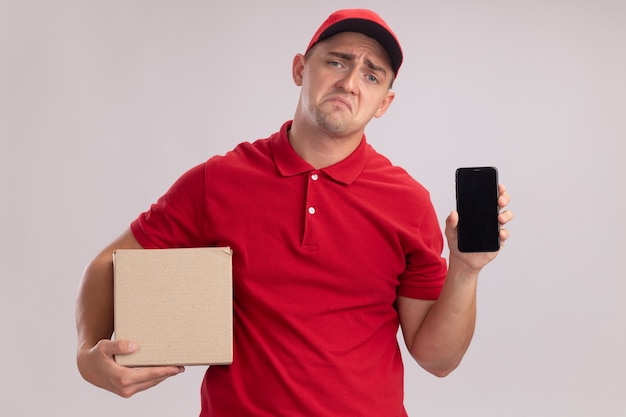 Smutny młody człowiek dostawy ubrany w mundur z czapką trzymającą pudełko i telefon na białym tle na białej ścianie