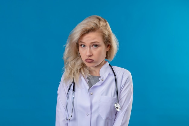 Bezpłatne zdjęcie smutny młody lekarz ubrany w stetoskop w sukni medycznej na niebieskiej ścianie
