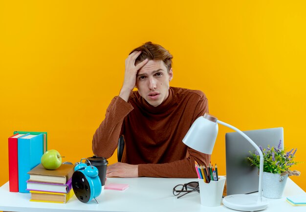 Smutny młody uczeń chłopiec siedzi przy biurku z narzędziami szkolnymi kładąc rękę na głowie na białym tle na żółtej ścianie