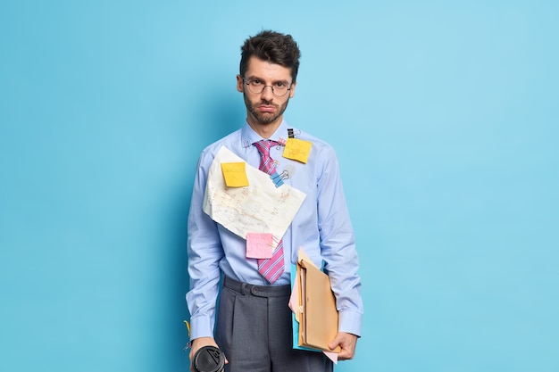 Smutny, nieogolony mężczyzna patrzy poważnie w kamerę trzyma kawę, a papiery zmęczony przygotowywaniem raportu ubrany w formalny strój przychodzi na spotkanie biznesowe