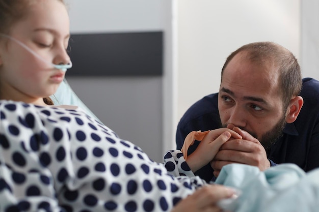 Bezpłatne zdjęcie smutny niespokojny opiekuńczy ojciec trzymający rękę śpiące chore małe dzieci w oddziale pacjenta szpitala pediatrycznego. chora dziewczyna za pomocą rurki nosowej w szpitalnym łóżku, podczas gdy uważny rodzic patrzy na nią.