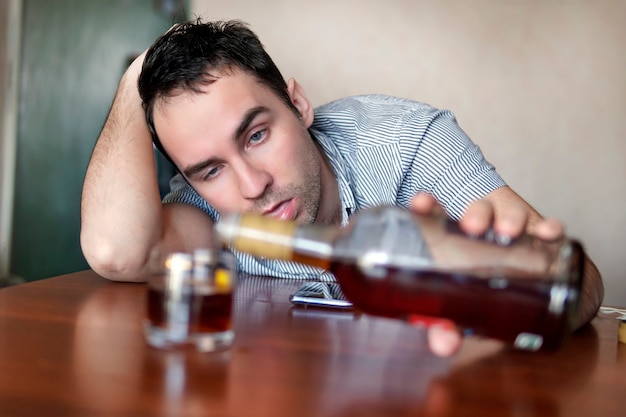 Smutny smutny młody alkoholik siedzi przy stole z butelką rumu. twarz pijanego mężczyzny. głupie emocje. depresja i alkoholizm.