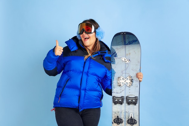 Snowboarding. Portret kobiety kaukaski na niebieskiej przestrzeni. Piękne modelki w ciepłych ubraniach