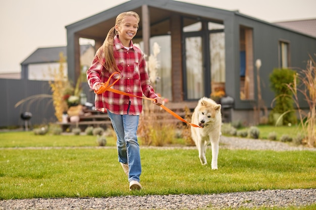 Bezpłatne zdjęcie spacery z psem. szczęśliwa urocza długowłosa dziewczyna w wieku szkolnym w zwykłych ubraniach biegająca z psem shiba inu na smyczy na trawniku w pobliżu wiejskiego domu w pogodny dzień