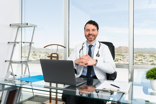Specjalista medyczny z wielką karierą. Przystojny lekarz uśmiechający się i nawiązujący kontakt wzrokowy siedząc za biurkiem w swoim gabinecie ze wspaniałym widokiem na miasto