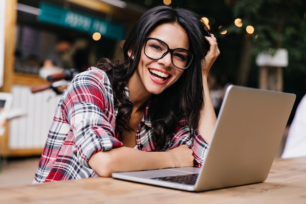 Spektakularna młoda kobieta o ciemnych włosach uśmiechnięta podczas pracy z komputerem. Zewnątrz zdjęcie emocjonalnej studentki w czarnych okularach.