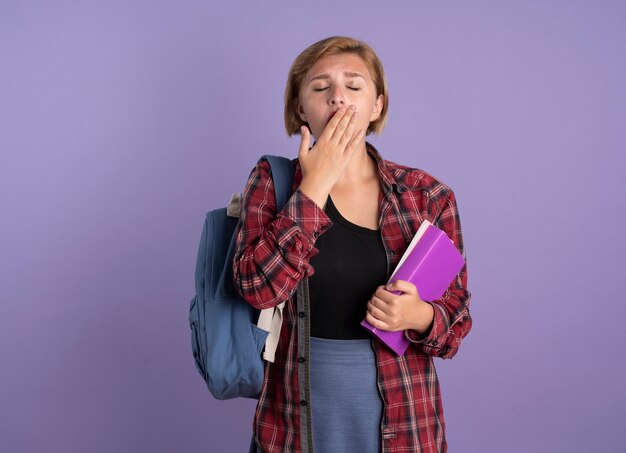 Śpiąca młoda słowiańska studentka w plecaku, kładąca rękę na ustach, trzymająca książkę i notatnik