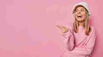 Bezpłatne zdjęcie spójrz na tę promocję szczęśliwa piękna kobieta śmieje się i wskazuje palec wskazujący na bok reklamuje produkt ma zadowolony wyraz nosi modną zimową panamę i sweter odizolowane na różowej ścianie studia