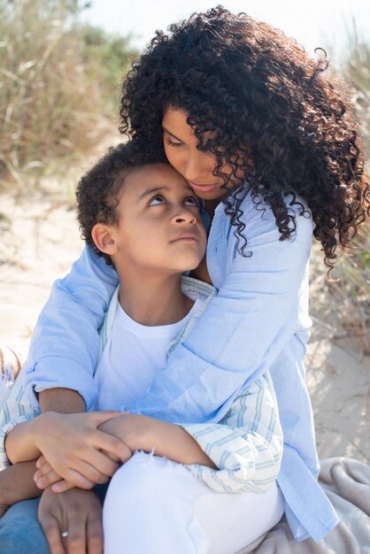Spokojna afroamerykańska matka i dziecko na plaży. Matka i syn w zwykłych ubraniach, siedząc na kocu, przytulając. Rodzina, relaks, koncepcja natury