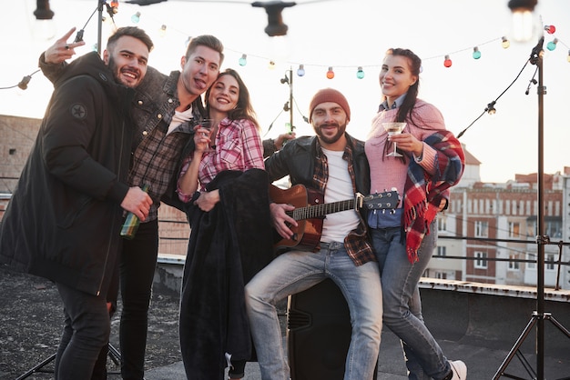 Spokojna i wesoła. Impreza na dachu. Pięciu dobrze wyglądających przyjaciół, którzy pozują do zdjęcia z alkoholem i gitarą