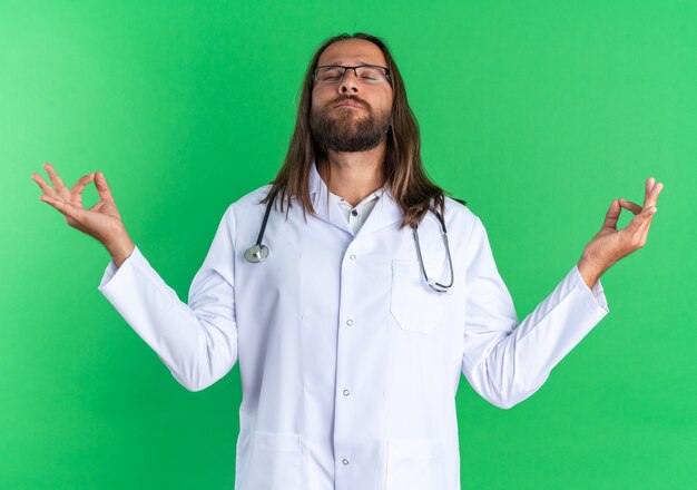 Spokojny dorosły lekarz-mężczyzna ubrany w szatę medyczną i stetoskop w okularach medytujący z zamkniętymi oczami odizolowanymi na zielonej ścianie