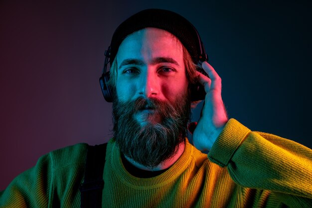 Spokojny, szczęśliwy, uśmiechnięty. Portret mężczyzny rasy kaukaskiej na tle gradientu studio w świetle neonu. Piękny męski model w stylu hipster w słuchawkach. Pojęcie ludzkich emocji, wyraz twarzy, reklama.