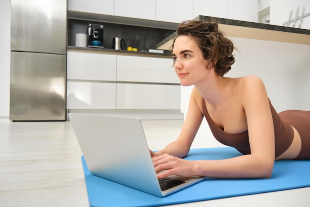 Bezpłatne zdjęcie sport i dobre samopoczucie dziewczyna fitness w odzieży sportowej leżącej na podłodze w kuchni gumowej macie do jogi za pomocą laptopa c