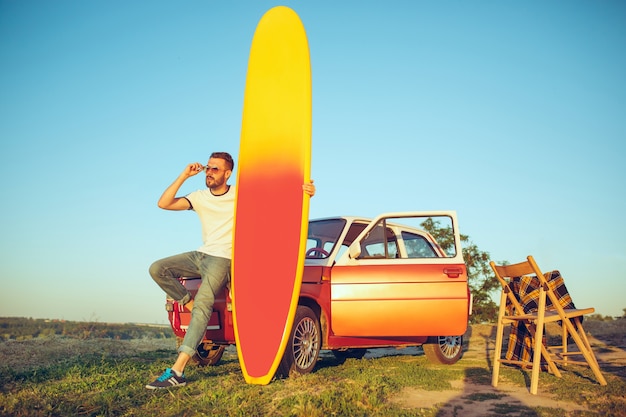 Sport, wakacje, podróże, koncepcja lato. Kaukaski mężczyzna stojący w pobliżu samochodu z deską surfingową w przyrodzie